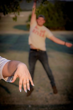 Woman showing off engagement ring with excited man celebrating in background outdoors.