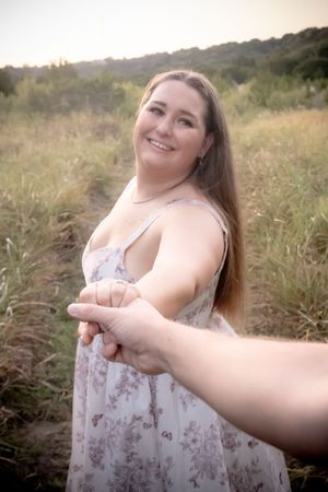 Smiling woman in floral sundress holds hands with someone in a sunny outdoor field at golden hour.