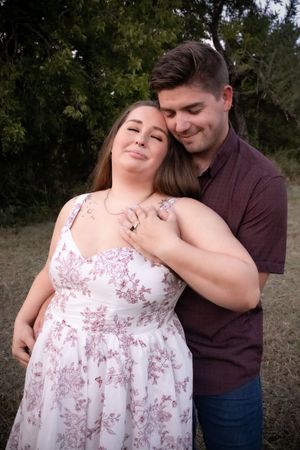 A couple embraces outdoors, woman in floral dress leans back into mans arms, both smiling softly.