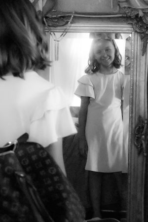 A black and white reflection in an ornate mirror shows someone wearing a white dress with flutter sleeves.