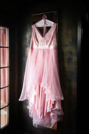 A pink wedding gown with sequined bodice and flowing tulle skirt hangs elegantly by a window in dramatic lighting.