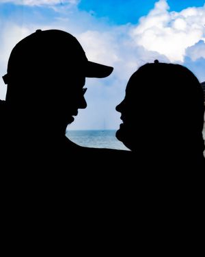 Silhouettes of two people with baseball caps against a bright blue sky with clouds and ocean background.