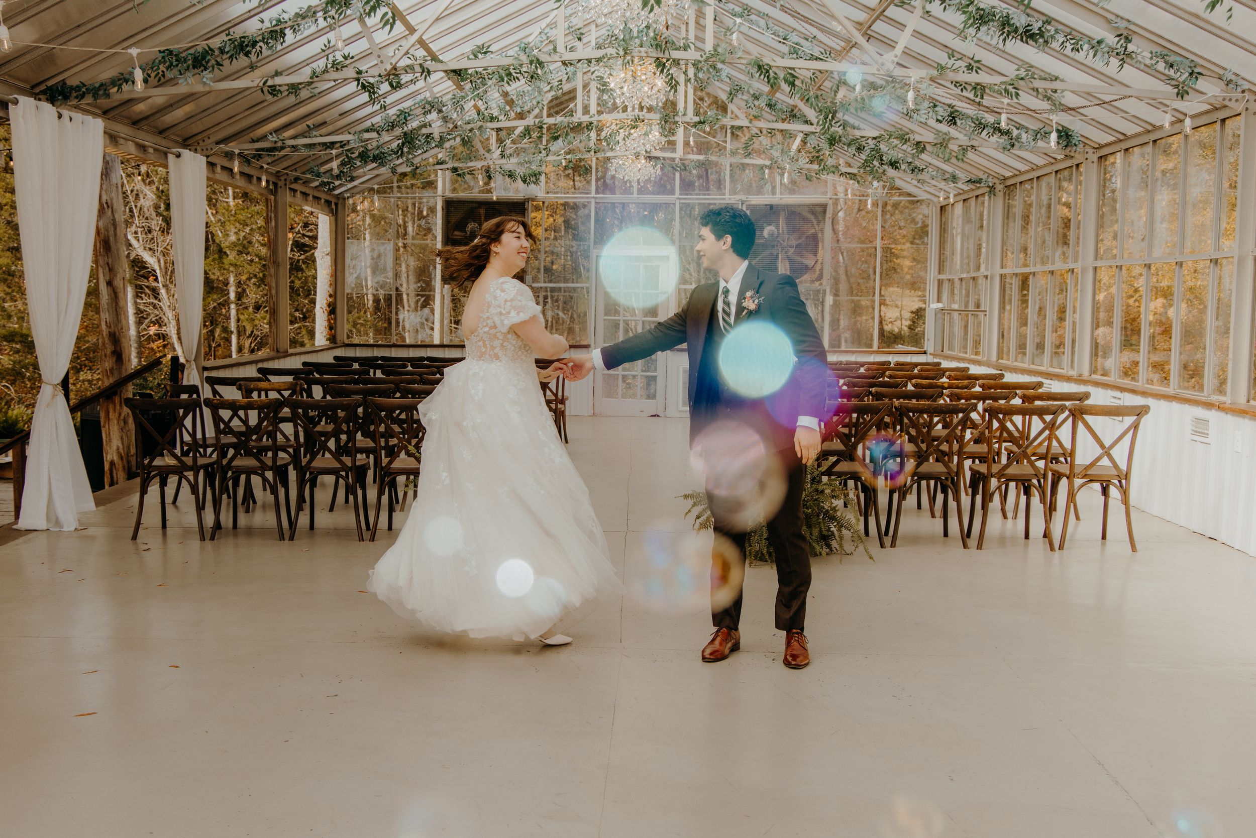 Wedding couple dances in a rustic reception venue with twinkle lights and wooden cross-back chairs.