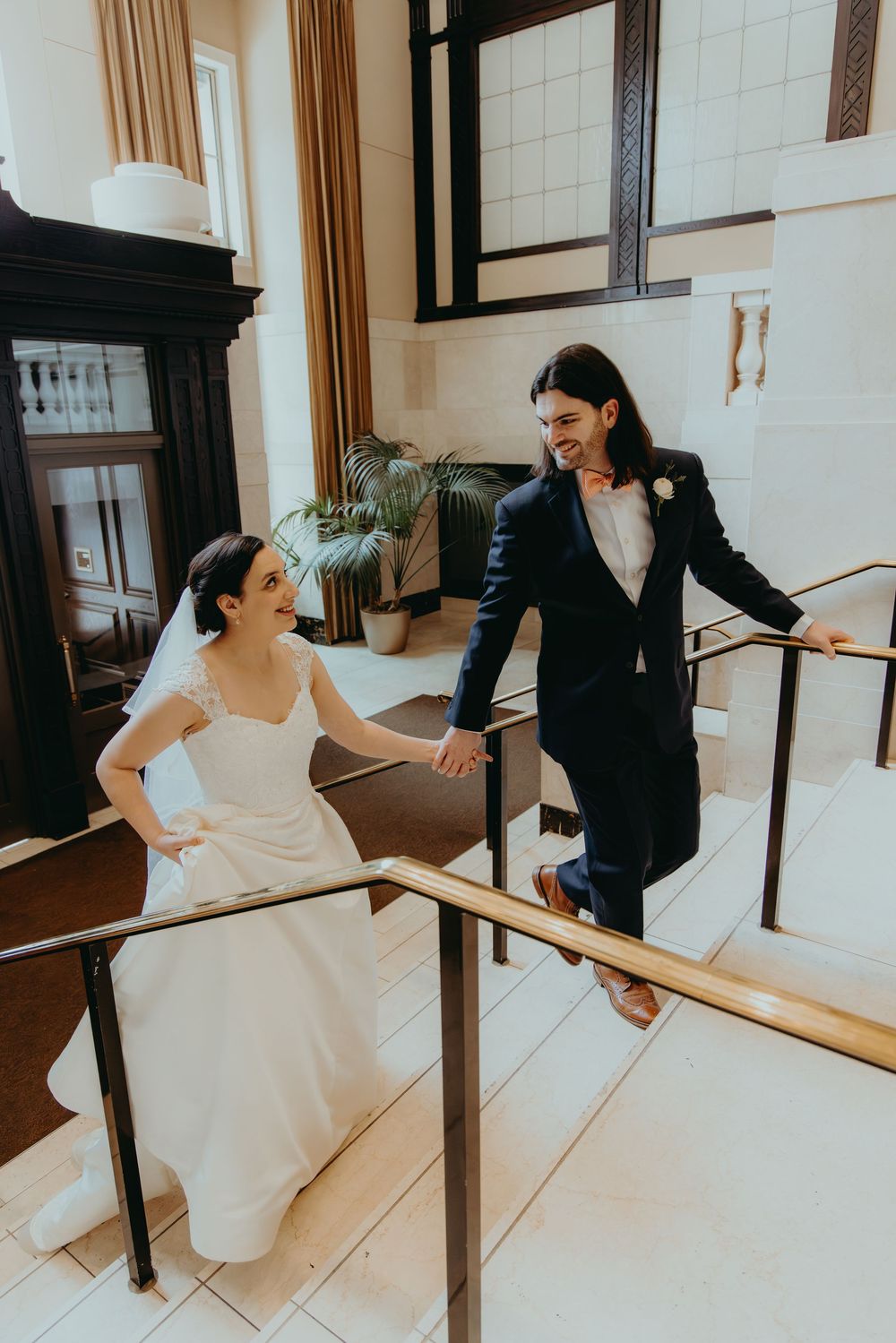 Smiling bride and groom holding hands on grand staircase in classic wedding venue.