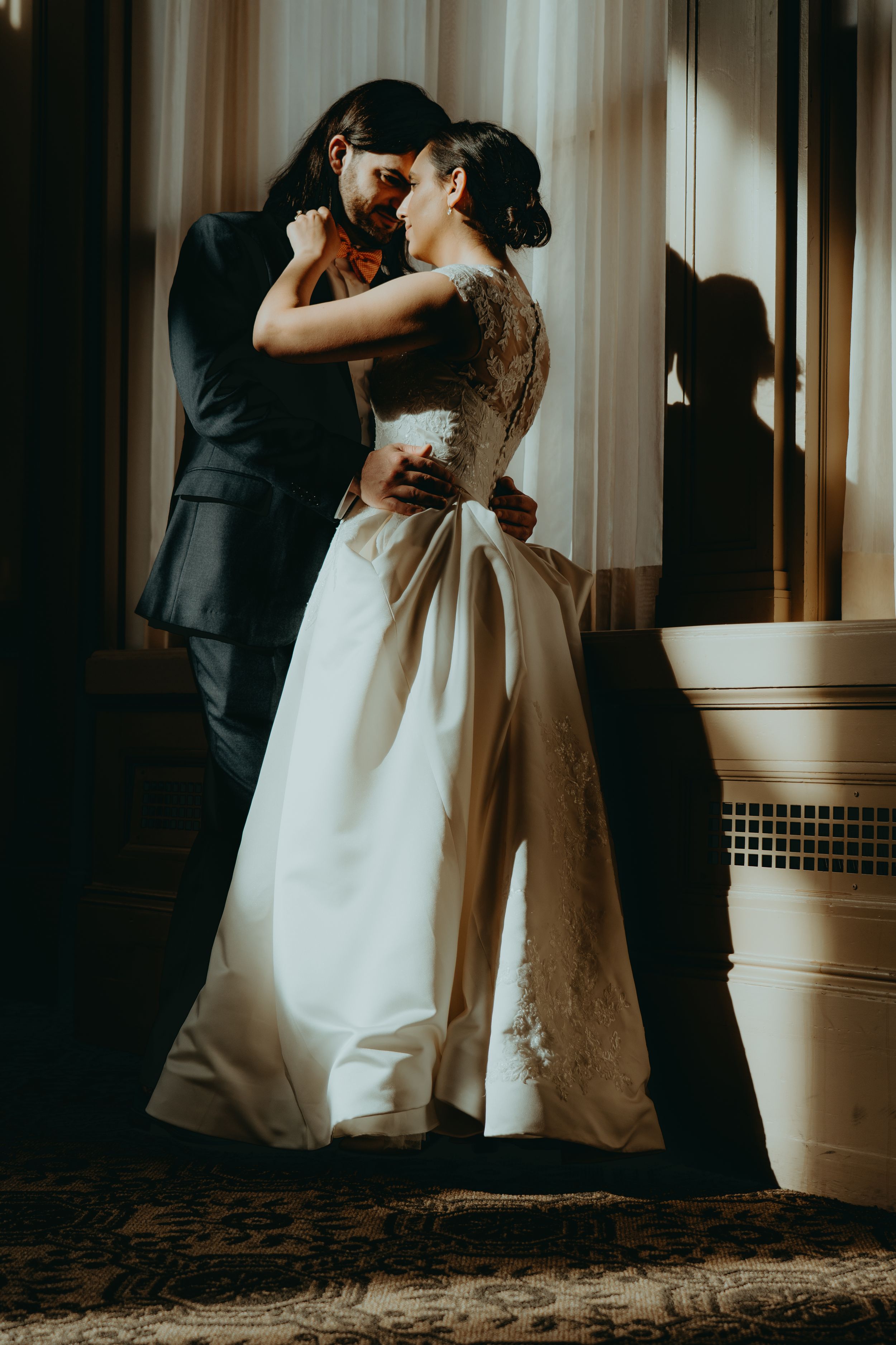 Bride and groom share a romantic embrace near a sunlit window, bride in elegant lace wedding gown.