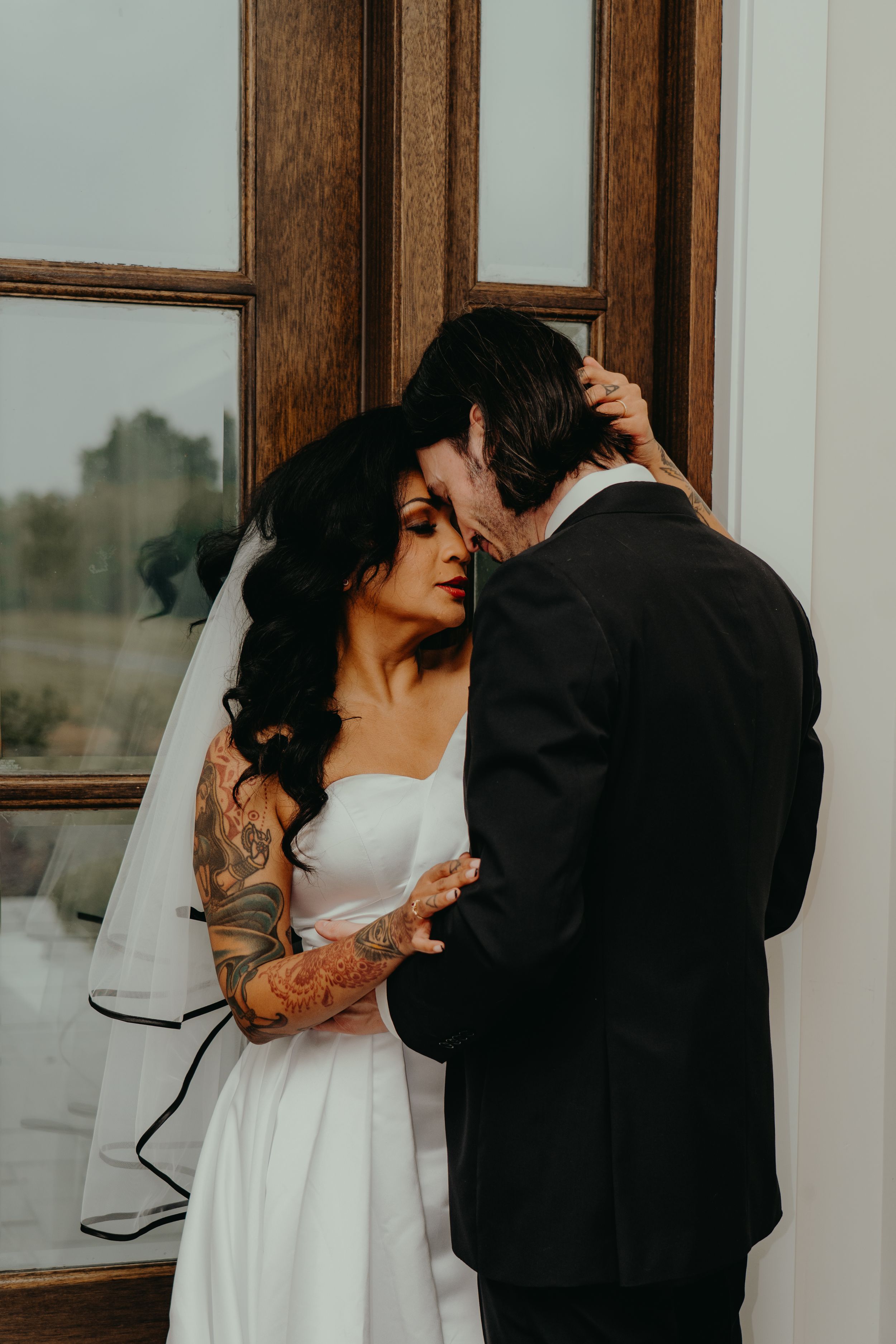 Tattooed bride in white gown and veil shares intimate moment with groom in black suit near wooden window.