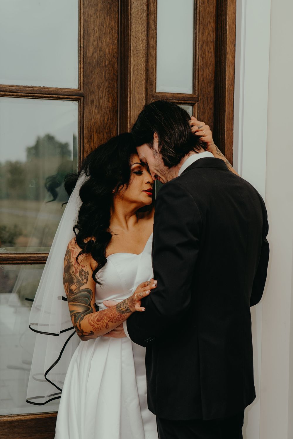 Tattooed bride in white gown and veil shares intimate moment with groom in black suit near wooden window.