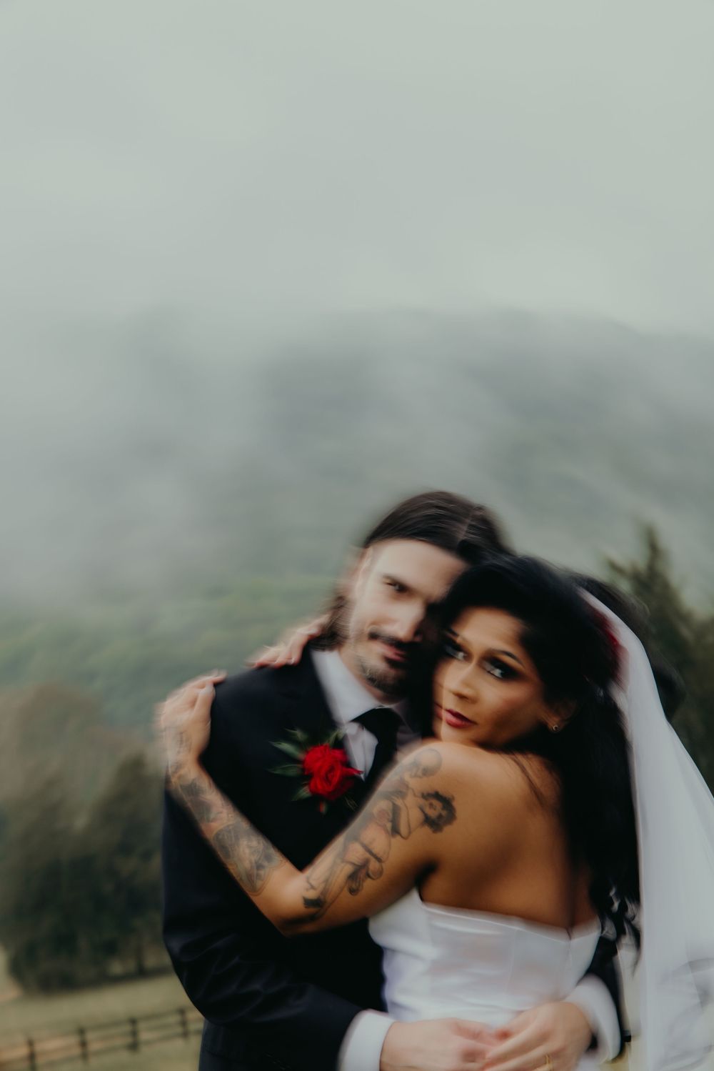 Tattooed bride and groom embrace in misty mountain wedding portrait, bride in white veil, groom in black suit with red boutonniere.