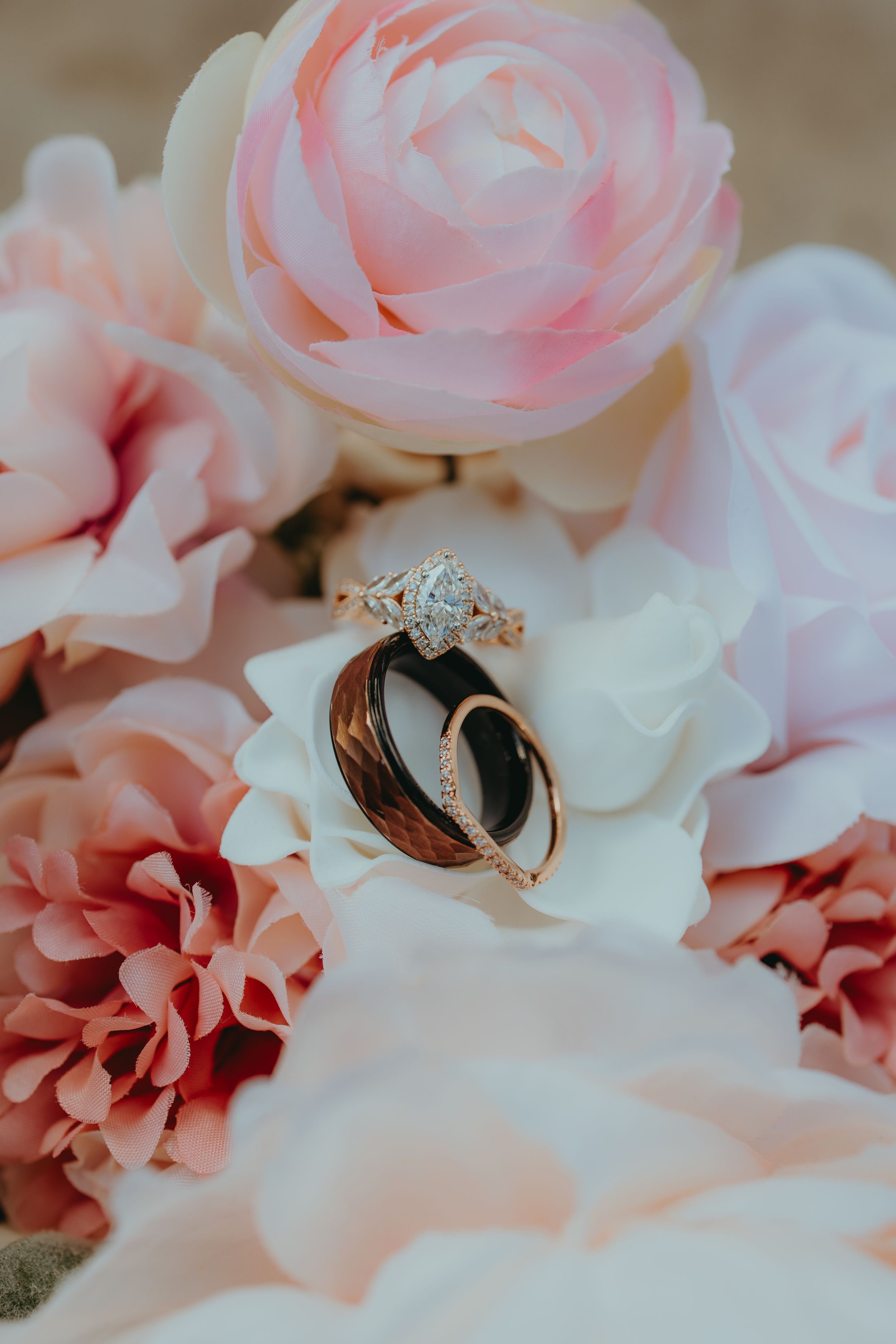 Wedding rings nestled among pink roses and white flowers, featuring a diamond engagement ring and dark wood band.