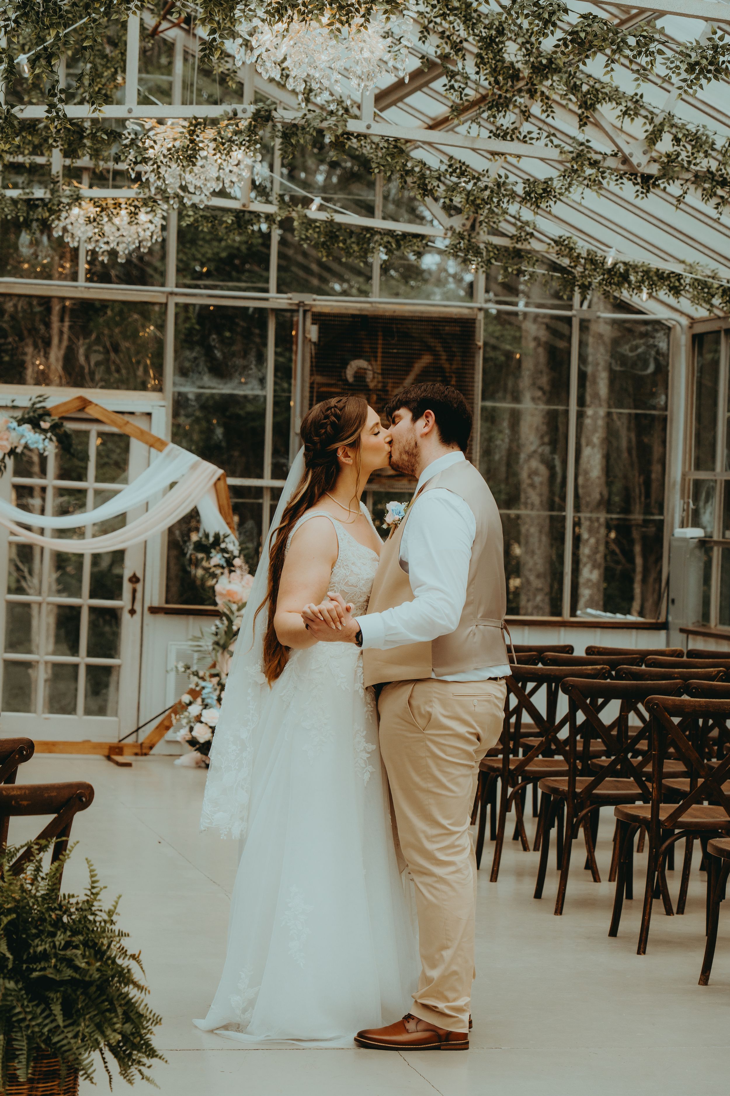 A bride and groom share their first kiss in a stunning glass greenhouse wedding venue adorned with chandeliers and greenery.