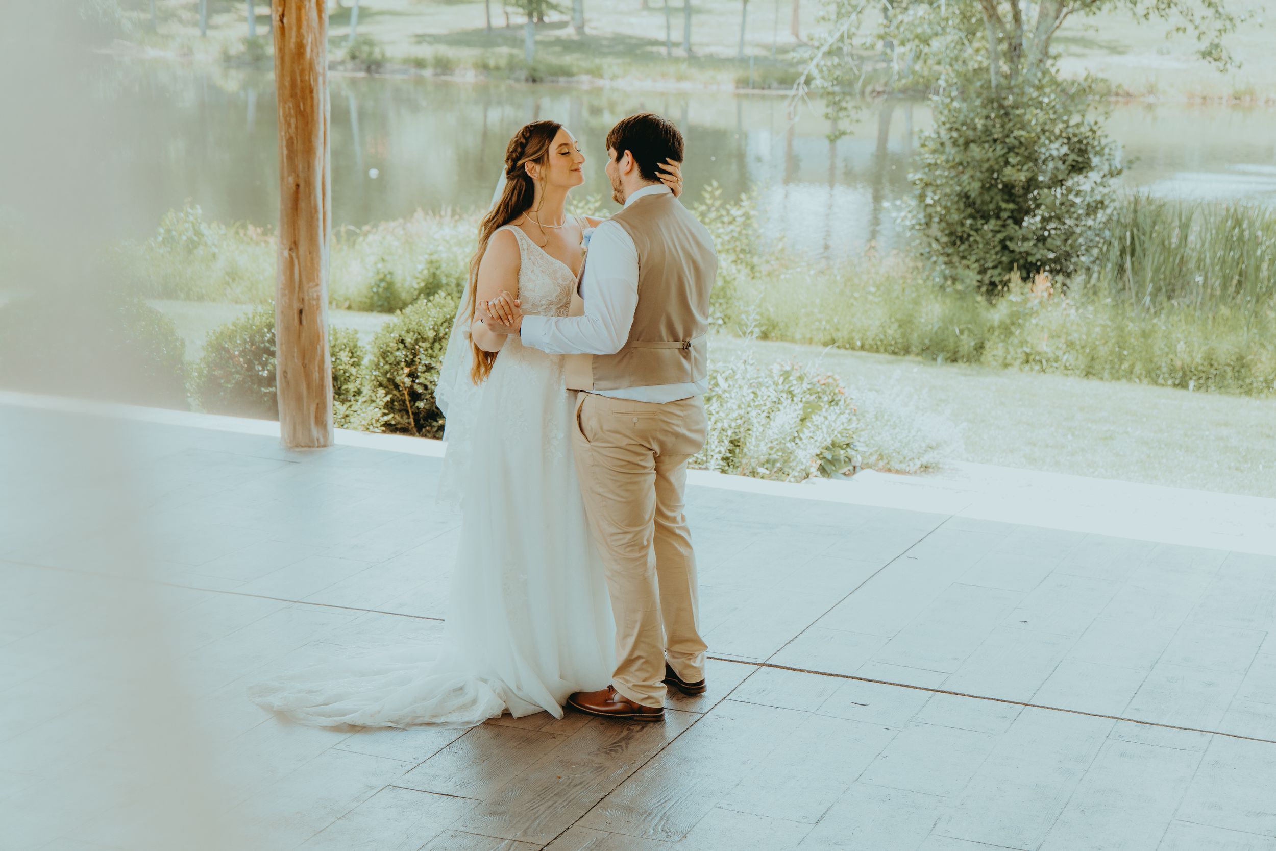 Bride and groom share a romantic moment in a sunlit venue with a scenic pond and greenery in the background.