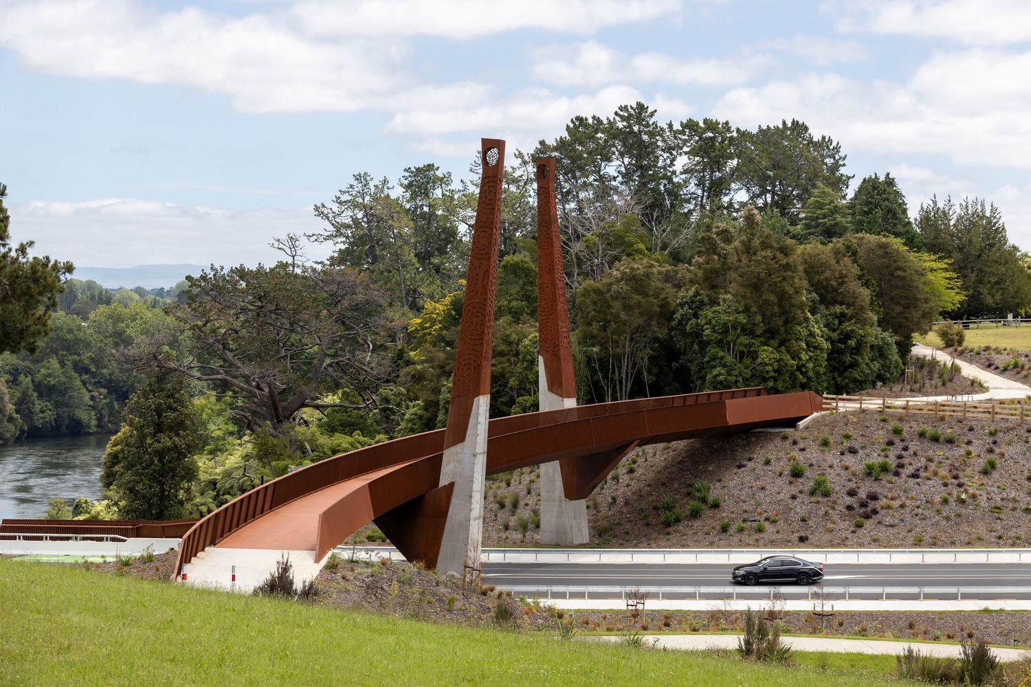 Sculptural pedestrian bridge with rusty red steel beams curves over a motorway and toward native bush.