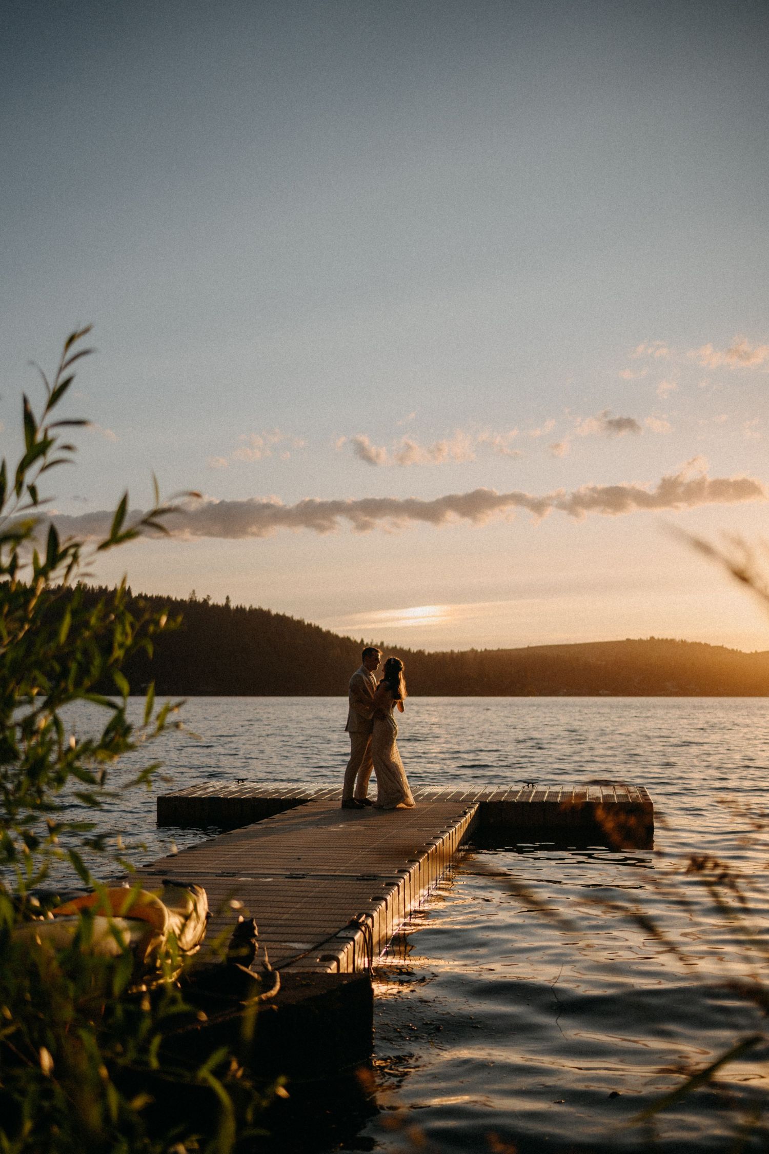 Silhouetted couple sharing romantic moment on lakeside dock during golden sunset with mountains in background.