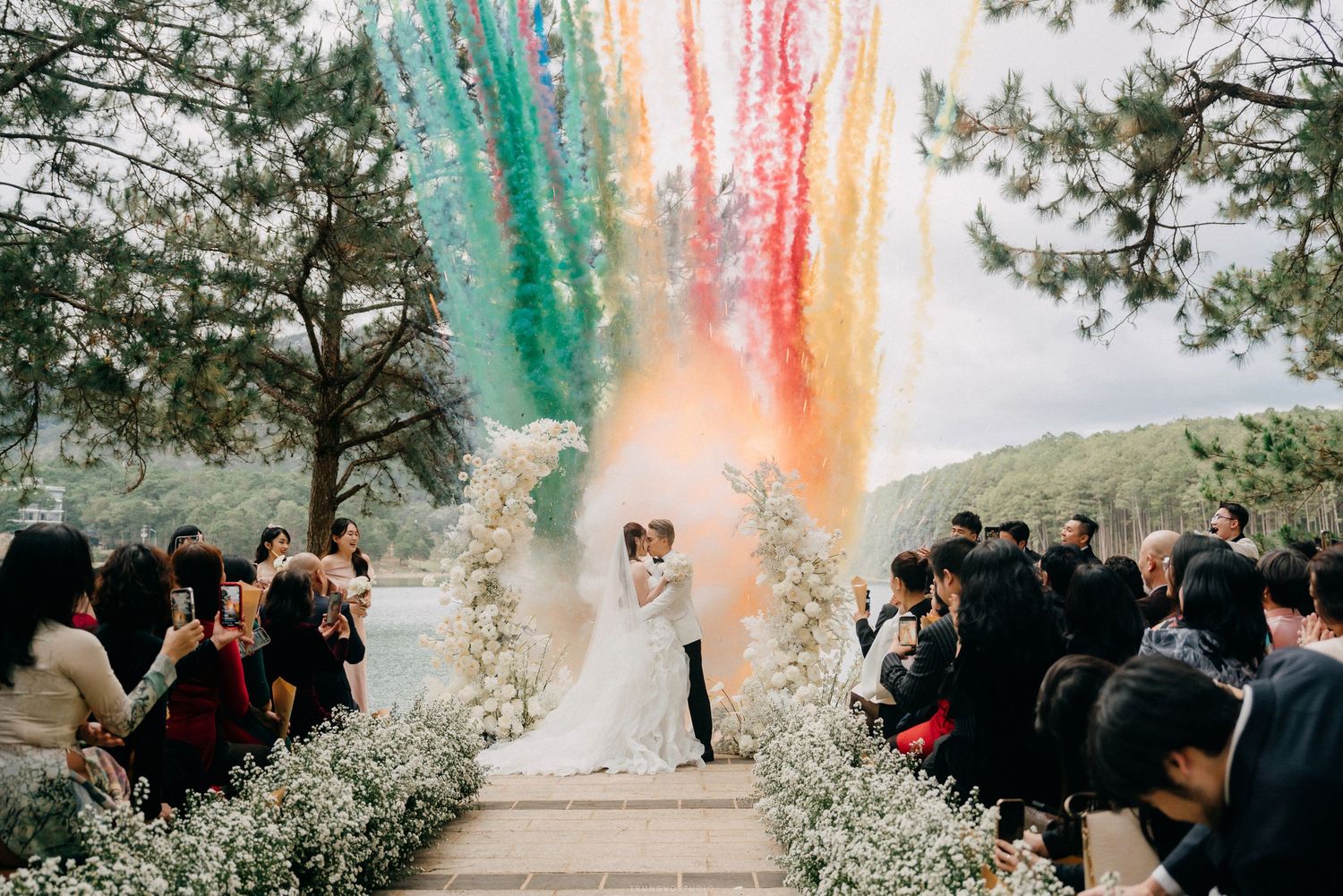 Couple kisses at outdoor wedding altar as colorful rainbow smoke cannons burst into sky above floral arch.