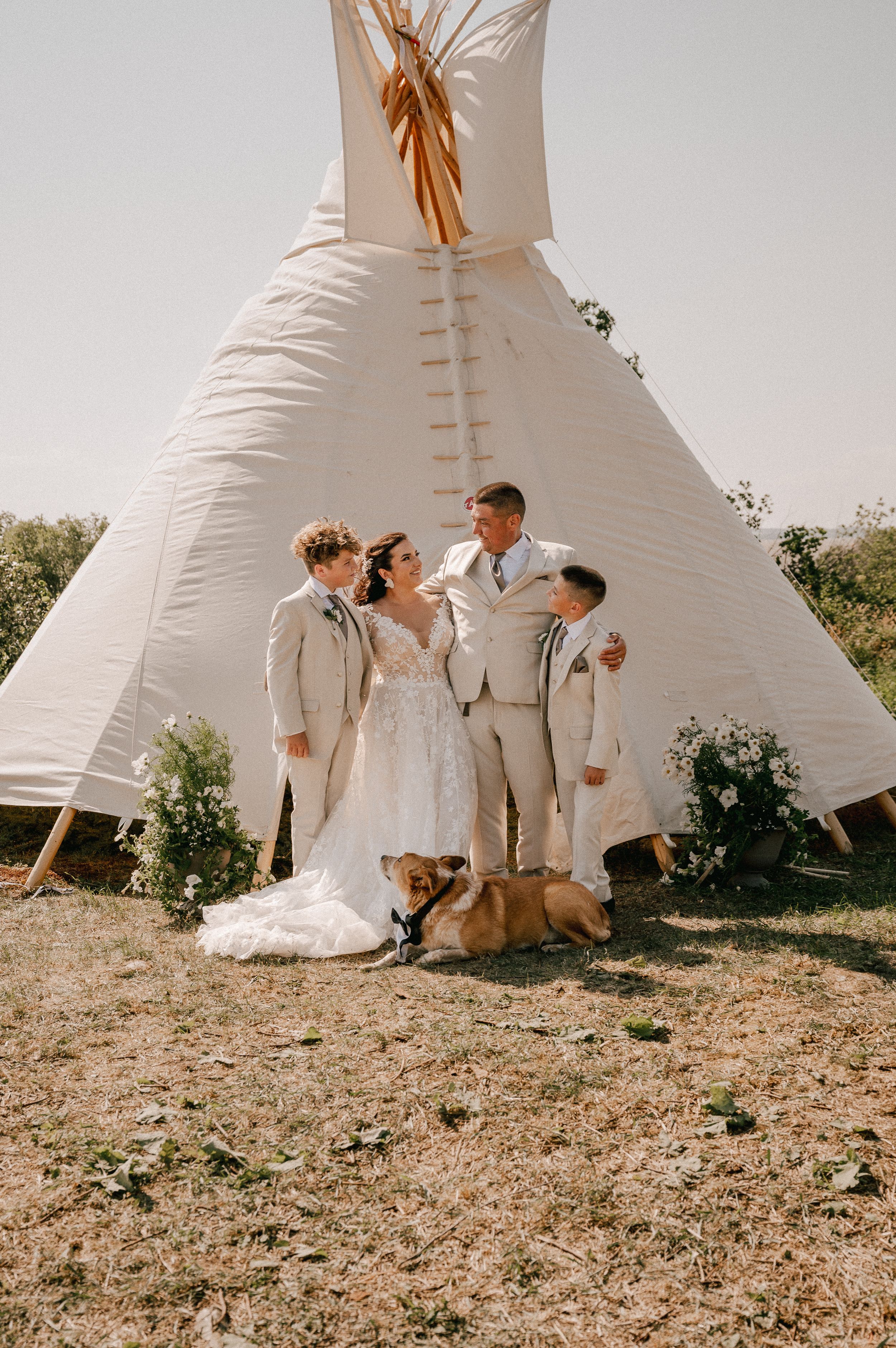Wedding party dressed in light colors poses in front of a white teepee on a sunny day with their dog.
