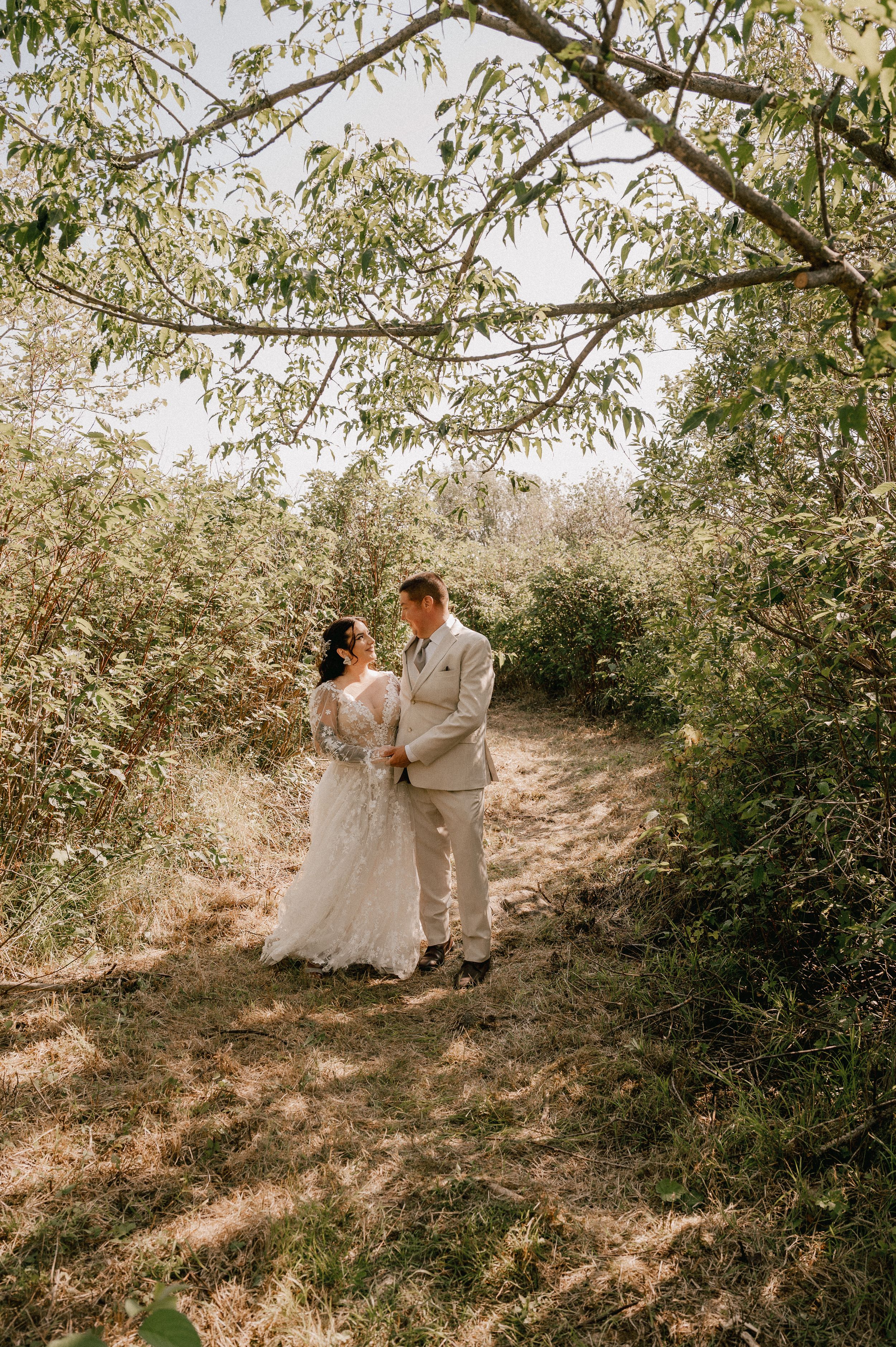 A couple in wedding attire gazes at each other while standing on a sunlit forest path surrounded by leafy trees.