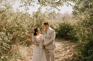 A couple in wedding attire holds hands on a sunlit path surrounded by leafy green foliage.