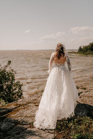 A white lace wedding dress silhouetted against a waterfront view on rocky terrain.