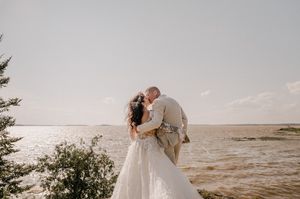 Romantic wedding couple embraces on rocky coastal cliff at sunset with ocean waves in background.