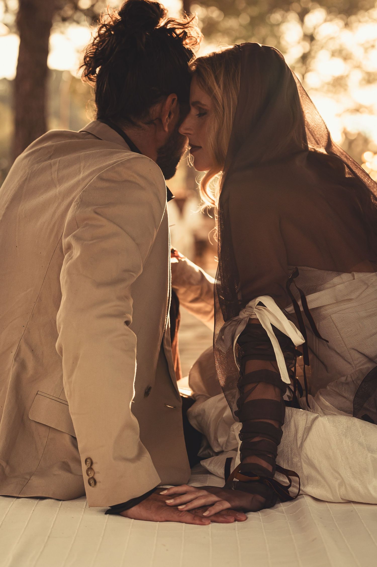 A romantic couple in light-colored clothing share an intimate moment at sunset with warm golden backlighting.