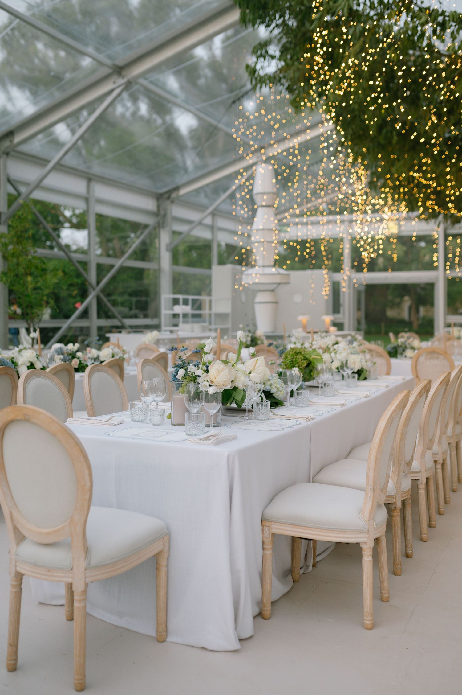 Elegant wedding reception table set in a glass greenhouse with white linens, cream chairs and twinkling lights overhead.