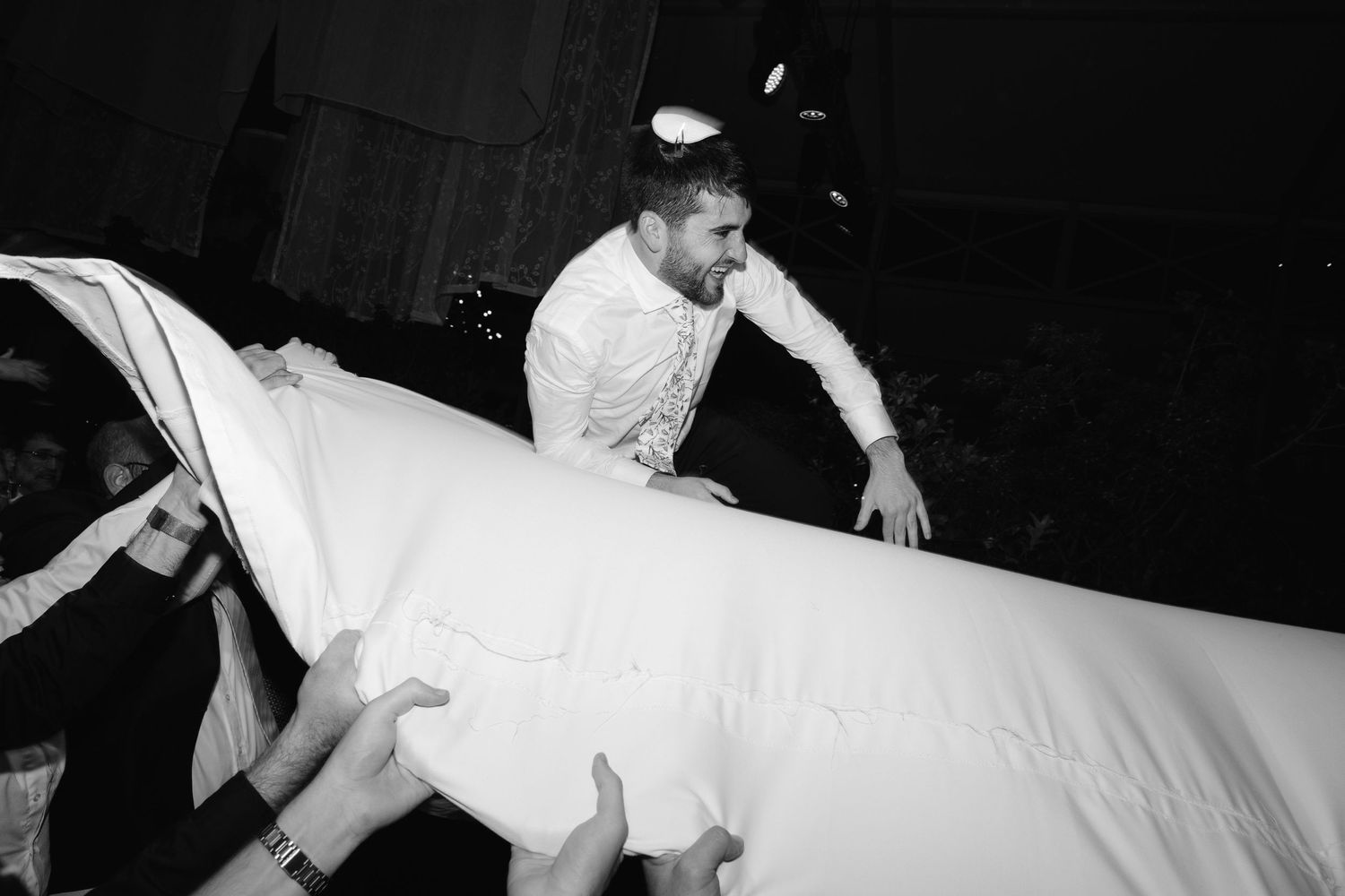 Black and white photo of wedding guests lifting and carrying a white tablecloth during a celebration.
