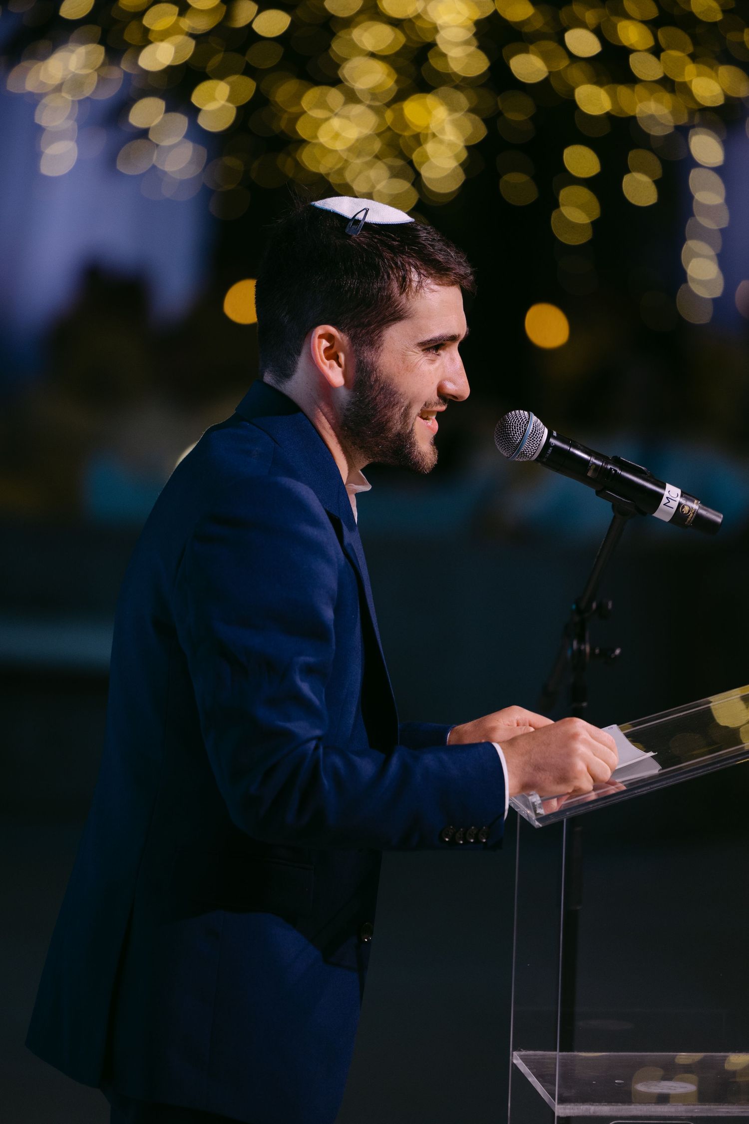 Speaker at podium delivering speech with bokeh lights in background during evening event.