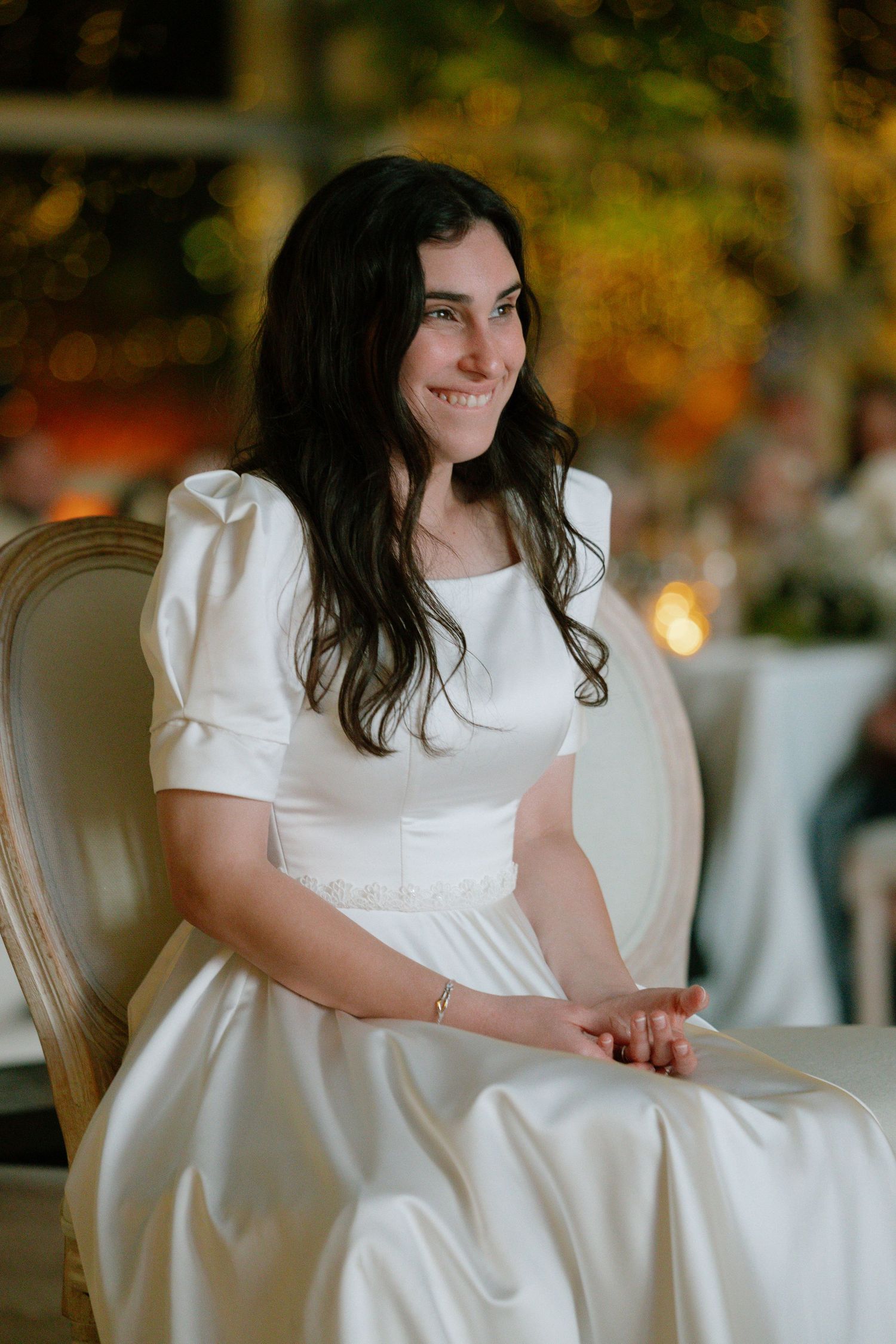 Bride in white dress seated elegantly at wedding reception with warm lighting in background.