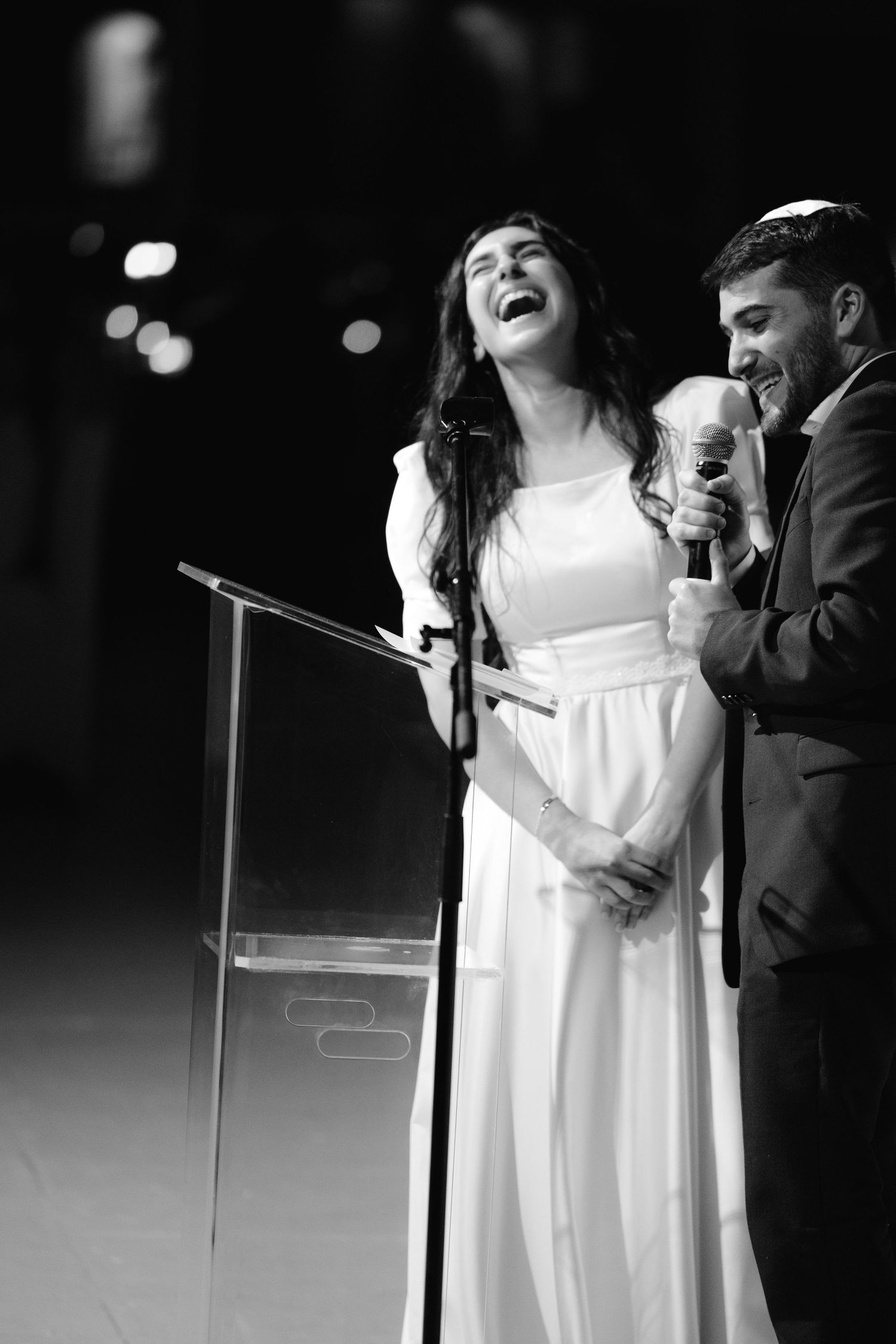 Black and white photo of a bride at a microphone during wedding reception speeches.