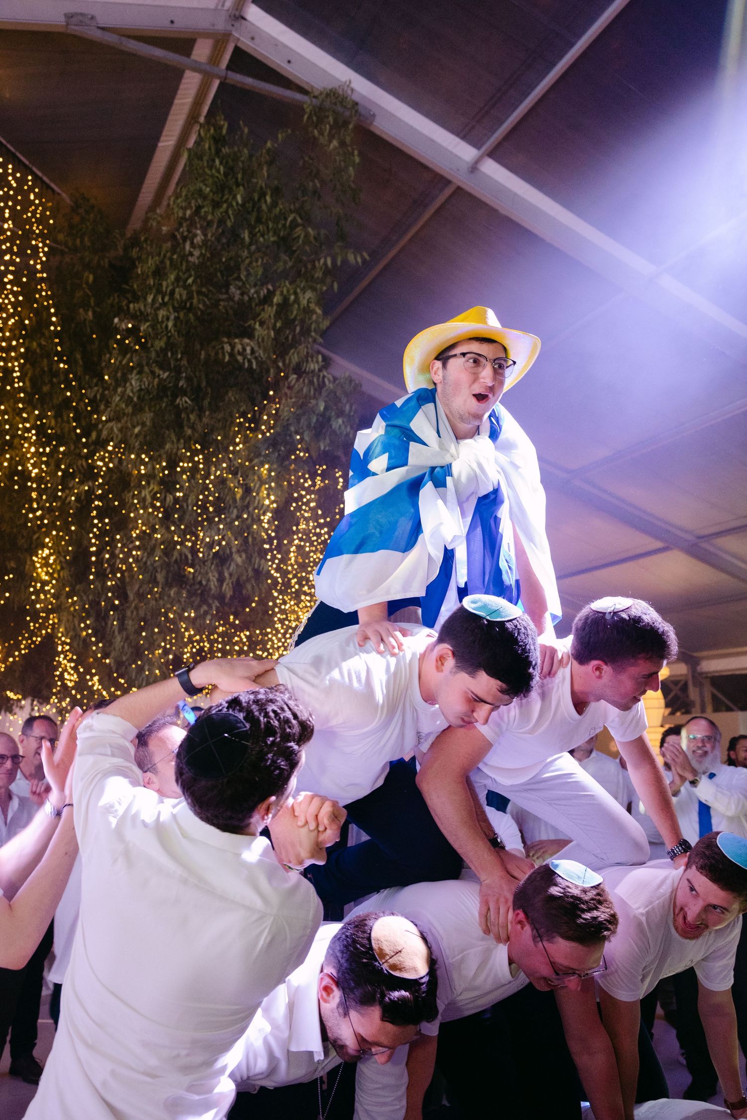 Energetic wedding guests dancing and jumping in celebration at a reception with twinkle lights in the background.