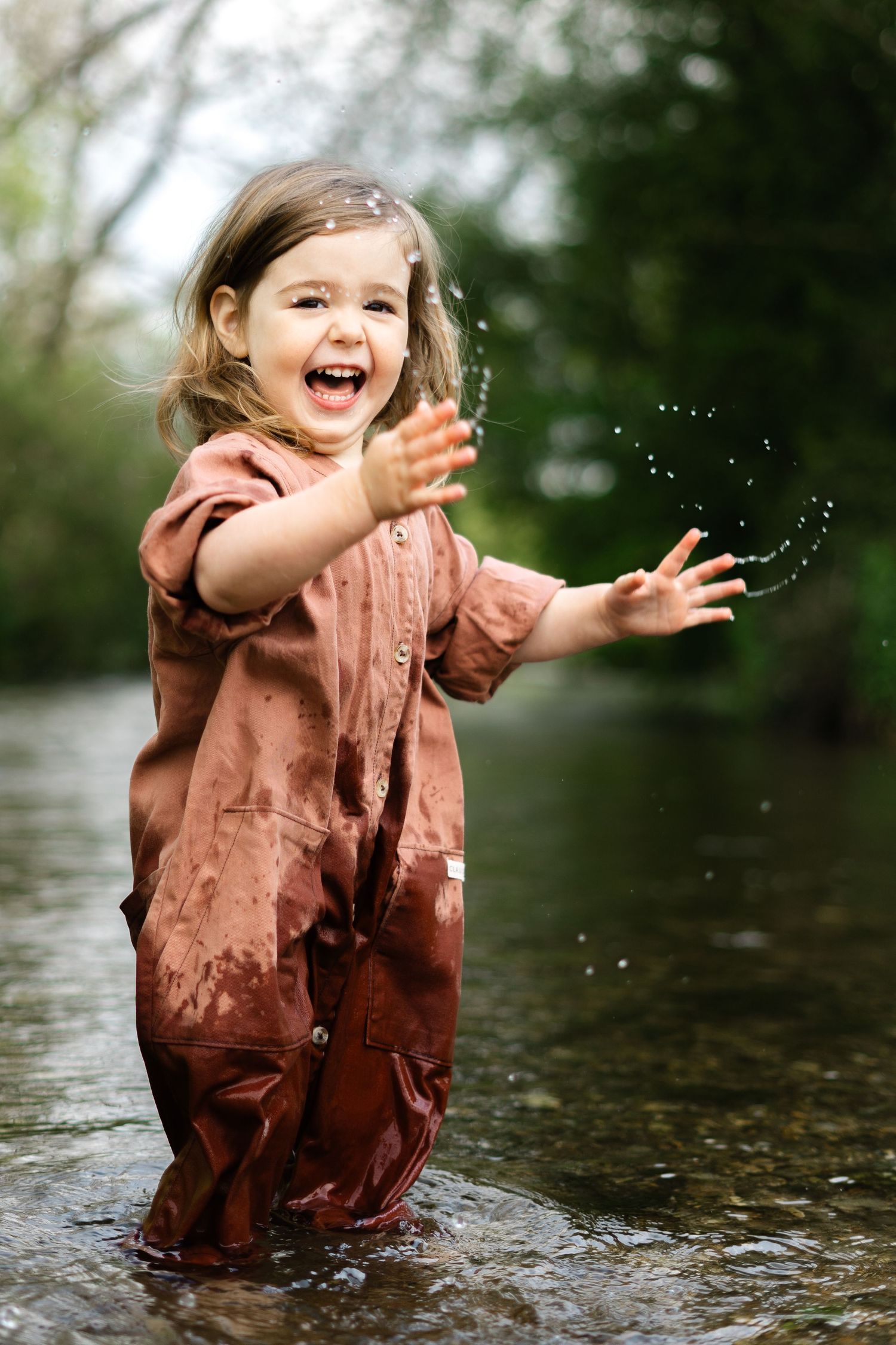 A joyful child in a brown dress splashes and plays in shallow water during an outdoor photoshoot.