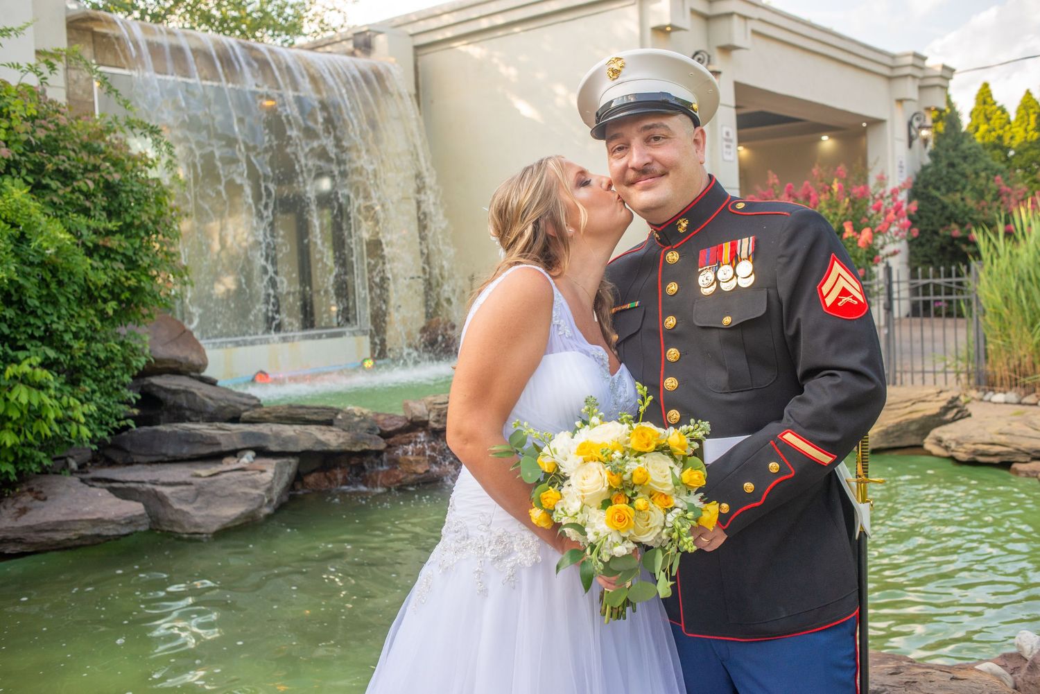 A couple in wedding attire embraces near a waterfront garden with bright yellow sunflower bouquet and military dress uniform.