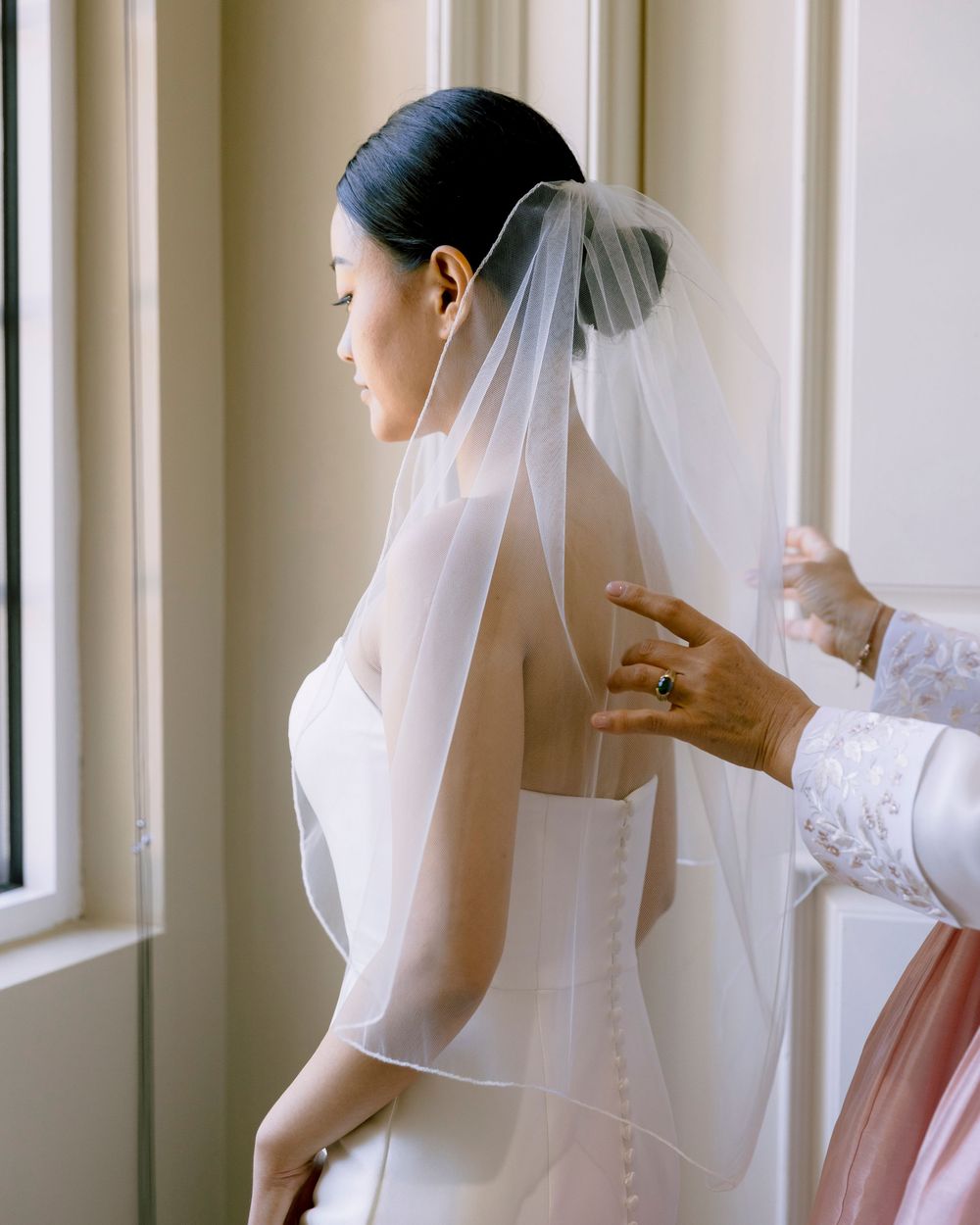 A bride in a white veil getting ready by a window with soft natural lighting.