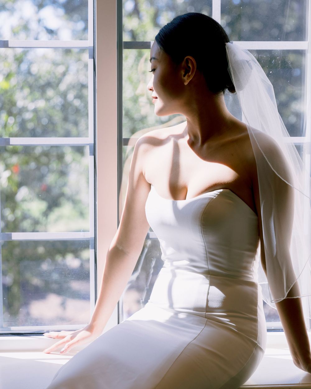 Bride in elegant white dress posing by bright window with natural light.