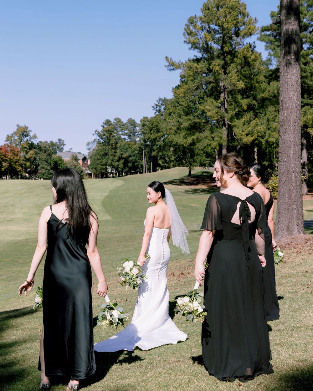 Wedding party walking across a grassy hill with bridesmaids in black dresses.