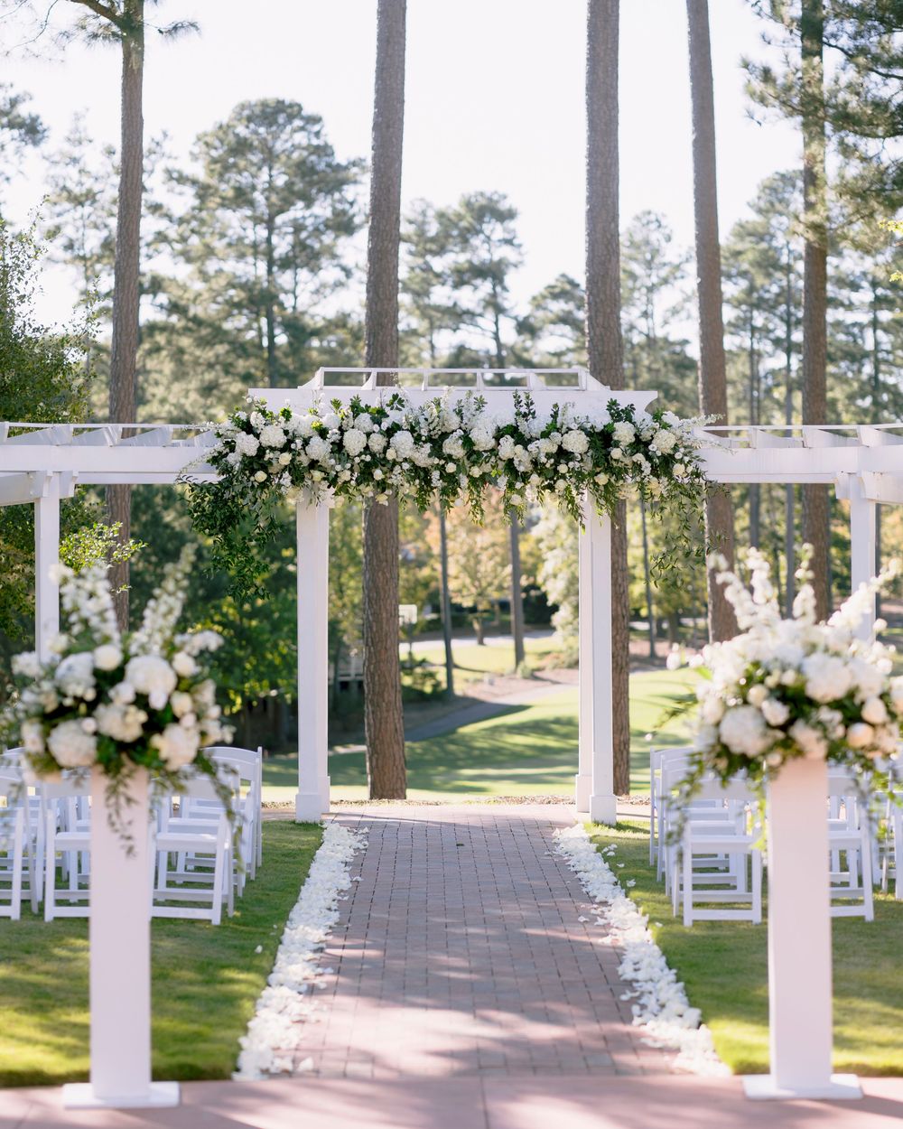 Outdoor wedding ceremony setup with white chairs and floral arch decorations under tall pine trees.