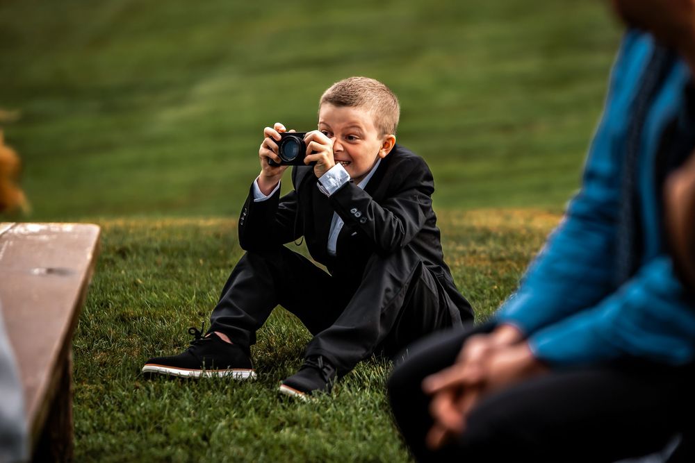A person in a blue jacket takes photos with a camera in a grassy outdoor setting.