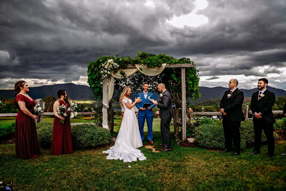 A dramatic wedding ceremony unfolds beneath dark storm clouds in a lush green field with mountains in the background.