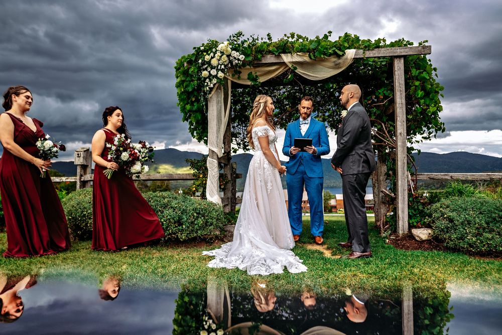 Outdoor wedding ceremony under wooden arch with dramatic stormy sky and mountain backdrop, reflected in water below.