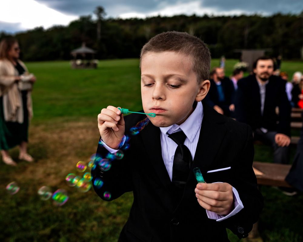 A young boy in formal attire plays with a bubble wand in a grassy field during an outdoor event.
