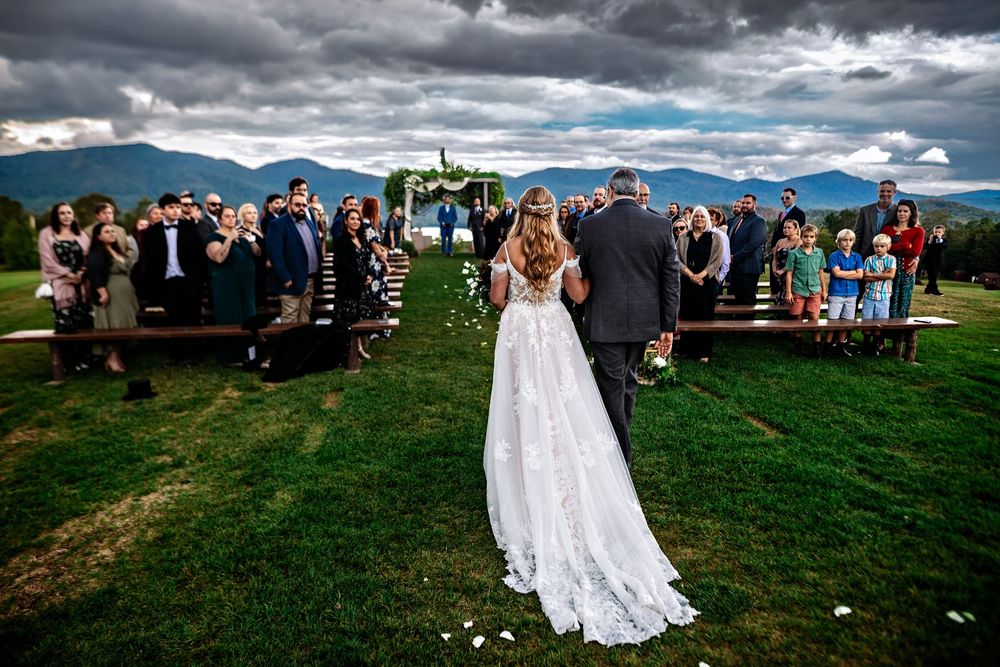 Panoramic mountain landscape serves as backdrop for an outdoor wedding ceremony with guests gathered on grassy hillside.