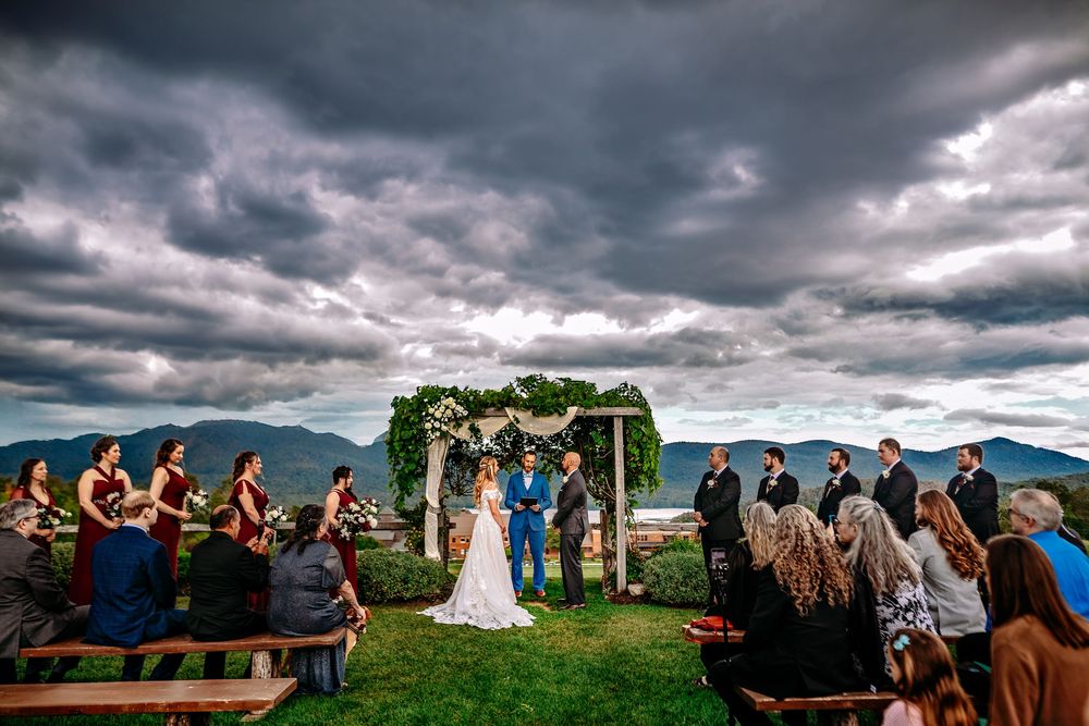 Dramatic cloudy skies frame an outdoor wedding ceremony with mountain views and floral arch decorations.