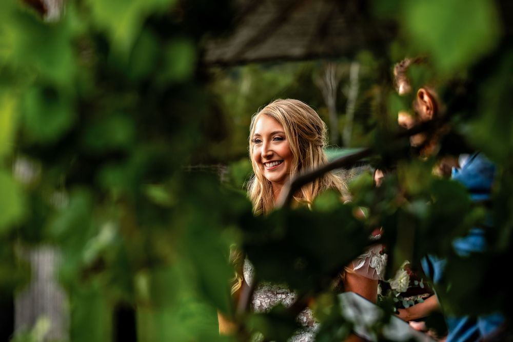 A person smiles while sitting in a lush green outdoor setting with sunlight filtering through the trees.