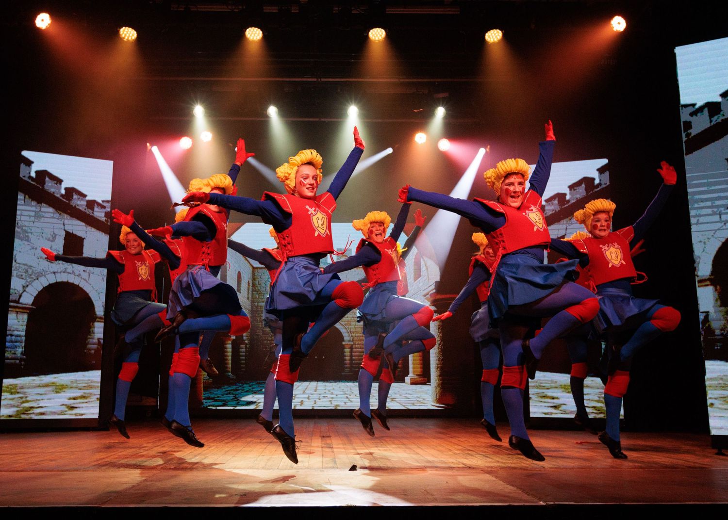 Dancers perform energetic choreography under warm red stage lights creating dynamic shadows and movement.