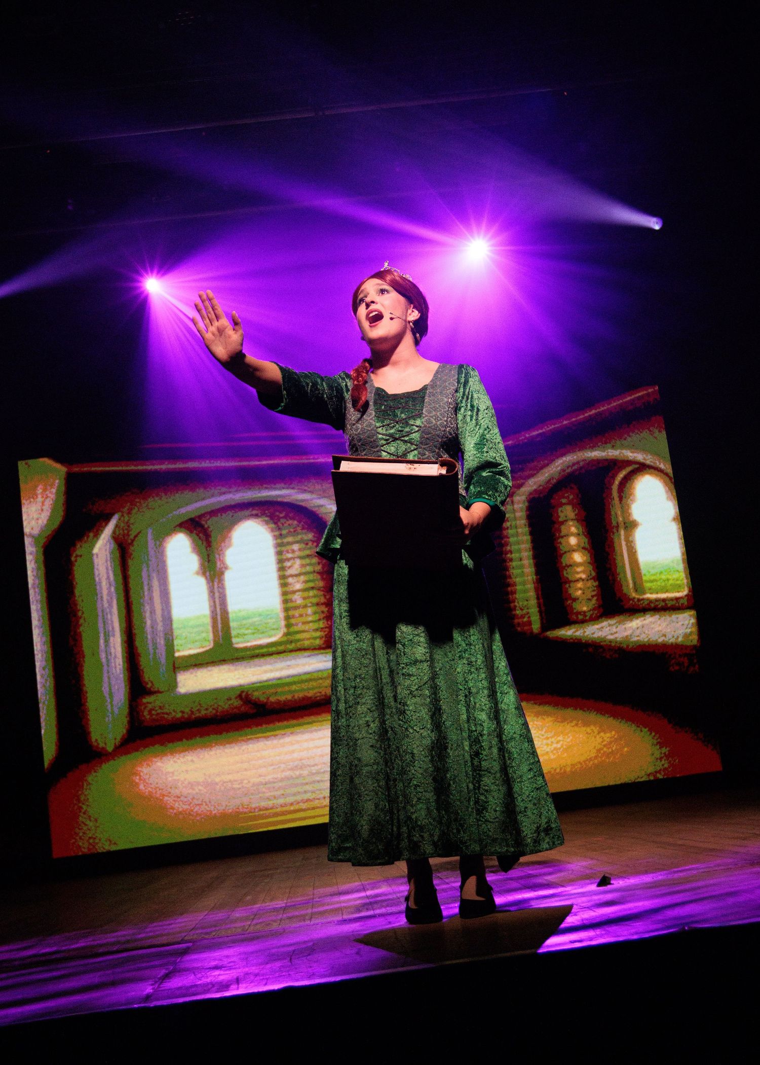 Opera performer in a green velvet dress gestures dramatically on stage under purple theatrical lighting.