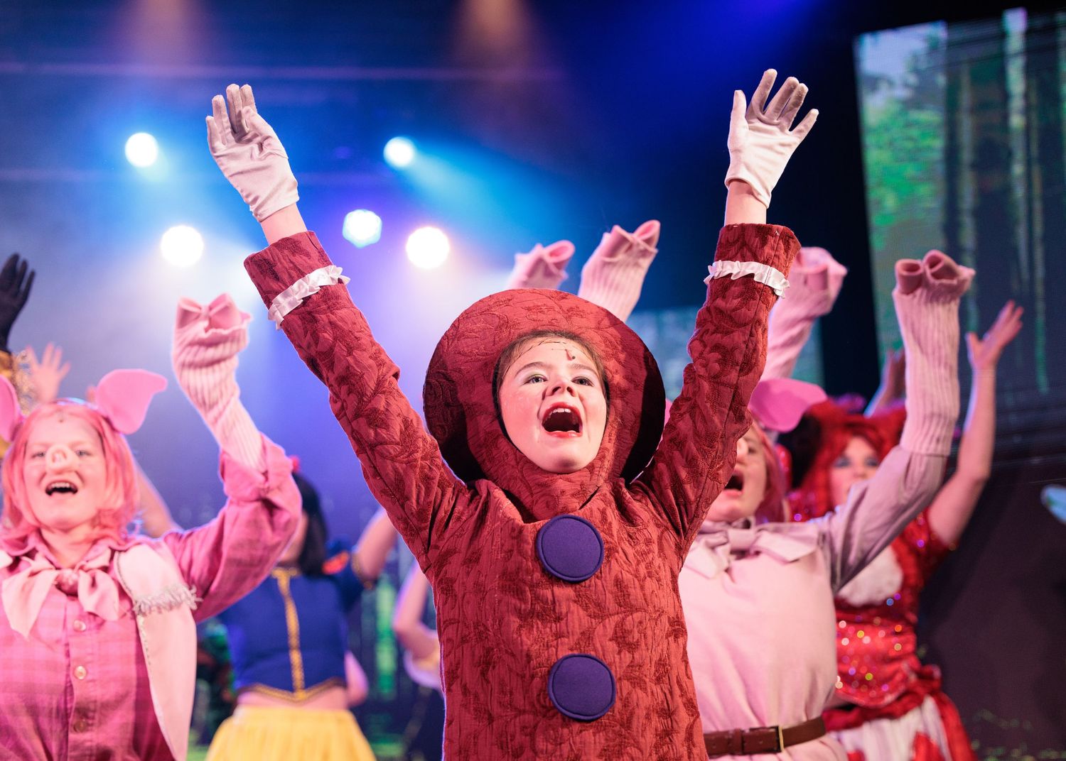 Theatrical performers in colorful costumes raise their arms in unison during an energetic stage performance.
