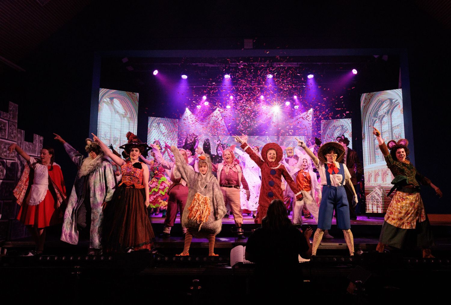 Wide shot of performers on stage during a theatrical production with dramatic purple and pink lighting effects.