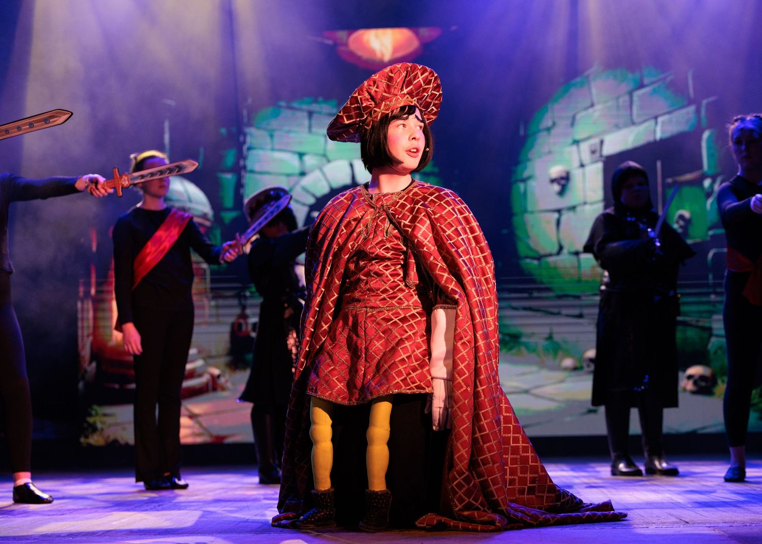 Theatrical performance showing characters in colorful costumes on stage with blue lighting and castle-like scenery.