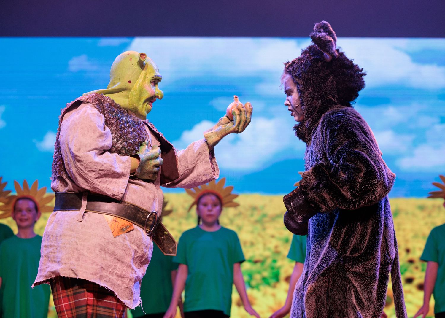 Actors performing in Shrek The Musical on stage with a bright blue sky and yellow field backdrop.