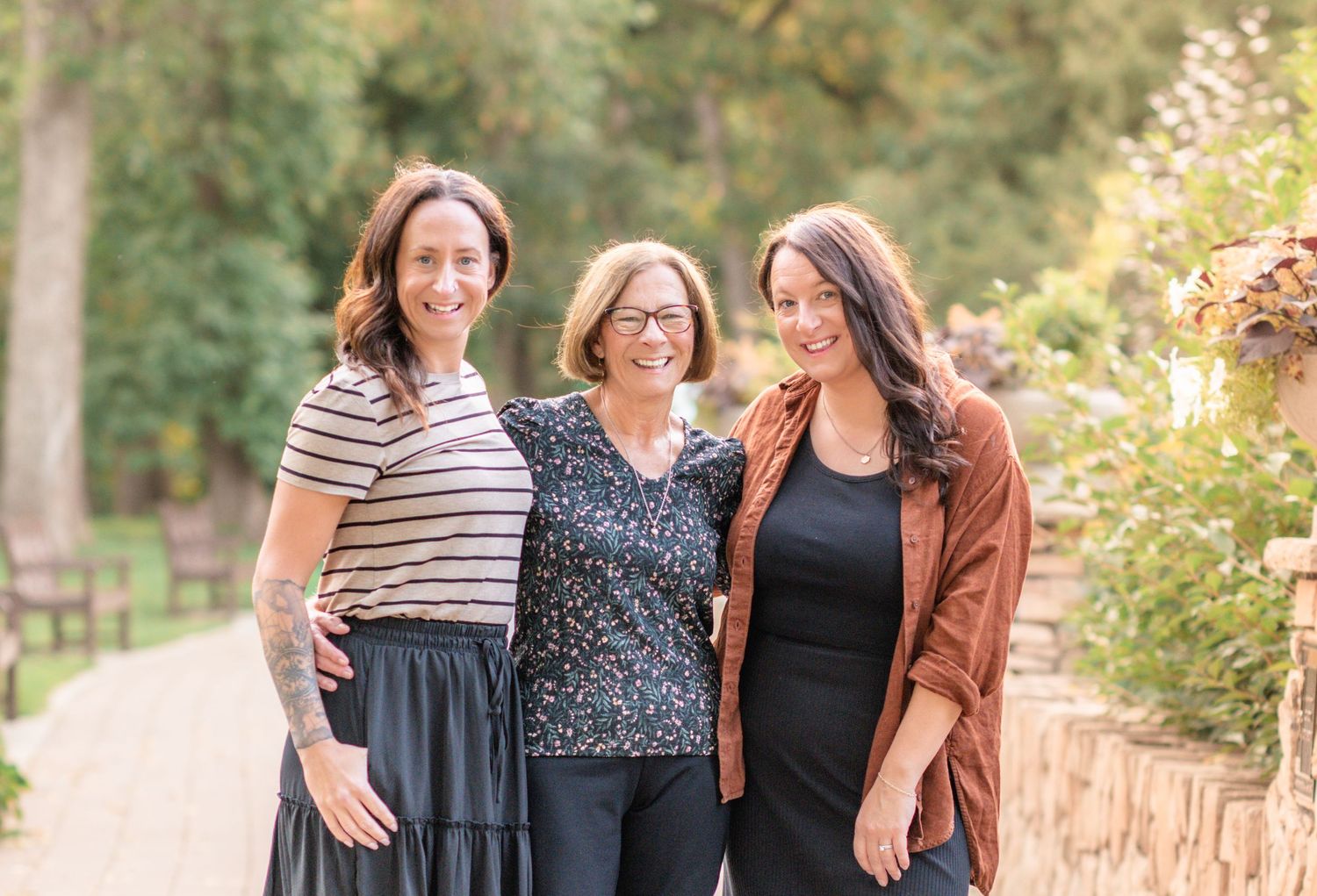 Three people stand together smiling on a garden path surrounded by greenery on a sunny day.